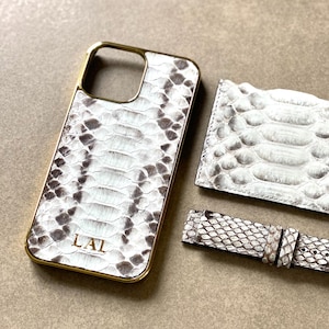 Pode incluir: Um conjunto de acessórios com padrão de pele de cobra em branco e castanho. O conjunto inclui uma capa de telemóvel com moldura dourada, um porta-cartões e uma bracelete de relógio. A capa do telemóvel tem as iniciais "LAL" em dourado.