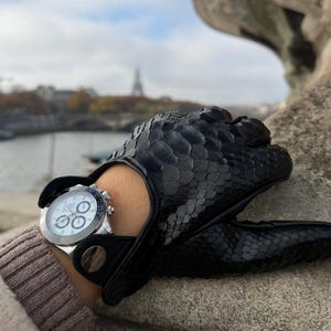 May include: Black leather driving gloves with a snakeskin pattern. A silver wristwatch with a white face and multiple dials is visible. The gloves are worn on a person's hand, with the Eiffel Tower in the background.