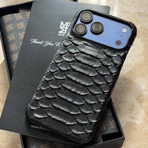 May include: Black textured phone case with a scale-like pattern, resting on a black box with the text "Thank You". The phone case has a blue camera module and is displayed with a matching patterned interior.
