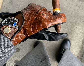 Puede incluir: Un cigarro marrón con una banda amarilla y naranja es sostenido por un guante de cuero marrón con un patrón de reptil. El guante tiene detalles en cuero negro. Se ve un reloj de pulsera con esfera azul. La persona lleva un abrigo gris y zapatos negros.