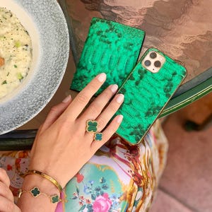 Könnte beinhalten: Ein grünes Handycase und eine Brieftasche mit Schlangenleder-Textur liegen auf einem Glastisch. Ein goldenes Armband und ein Ring mit grünen Kleeblatt-Akzenten sind zu sehen. Ein Teller mit Risotto und ein Getränk sind ebenfalls auf dem Tisch.