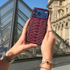 May include: A smartphone case with a deep burgundy, textured snakeskin pattern. The case features a dark blue camera module cutout with three circular lens openings and a small circular flash opening on the upper right side of the back panel.