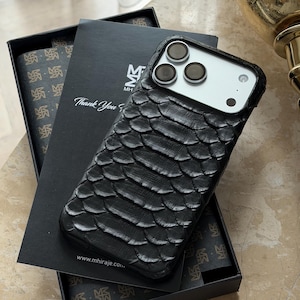 Könnte beinhalten: Schwarze, strukturierte Handyhülle mit schuppenartigem Muster, auf einer schwarzen Box. Die Hülle hat einen Kameraausschnitt und befindet sich neben einer Box mit dem Text "Thank You". Die Box hat ein sich wiederholendes Muster und die Website-Adresse www.mhiraje.com.