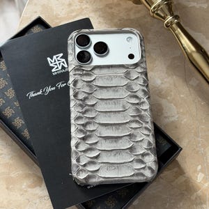 Puede incluir: Una funda gris para teléfono con textura de piel de serpiente y un recorte para una cámara de triple lente. La funda se muestra sobre una superficie de mármol junto a una caja negra que contiene una tarjeta con el texto "Thank You For" y un logotipo. Un objeto decorativo dorado está en el fondo.