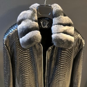 Może przedstawiać: Czarna skórzana kurtka z szarym kołnierzem z futra. Kurtka ma zamek błyskawiczny i wzór skóry węża.