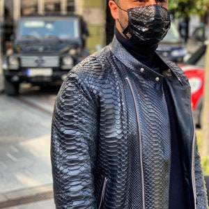 Pode incluir: Um homem vestindo uma jaqueta de couro preta com padrão de pele de cobra. Ele está vestindo uma camiseta de gola alta preta e uma máscara facial preta com padrão de pele de cobra.