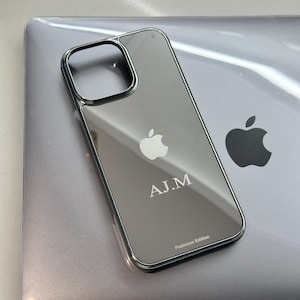 Puede incluir: Una funda transparente para tel&eacute;fono con marco plateado, apoyada sobre un port&aacute;til plateado. La funda tiene el logotipo de Apple y las iniciales "A.J.M" grabadas. Tambi&eacute;n es visible el texto "Platinum Edition".