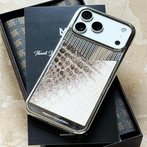 May include: A silver-framed phone case with a white and brown crocodile-skin pattern. The case is labeled "Platinum Croco Edition" and rests on a black box with a matching design. The phone case has three camera lenses.