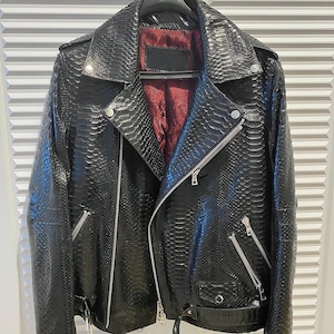 Pode incluir: Casaco de couro preto com padrão de pele de cobra, com gola entalhada, zíper assimétrico e ferragens prateadas. O casaco tem forro interior vermelho e detalhe de cinto na cintura. Está pendurado num cabide preto.