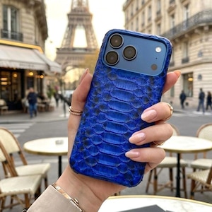 May include: A vibrant blue phone case with a snakeskin texture. The case has a camera module with three lenses. The background features a Parisian street scene with the Eiffel Tower in the distance.