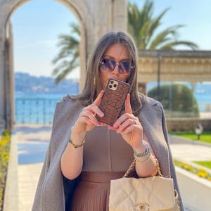 May include: A brown snakeskin-effect phone case is held up. The person is wearing sunglasses, a tan outfit, and carrying a cream quilted handbag with a gold chain strap. The background features a stone archway and palm trees, with a view of the sea.