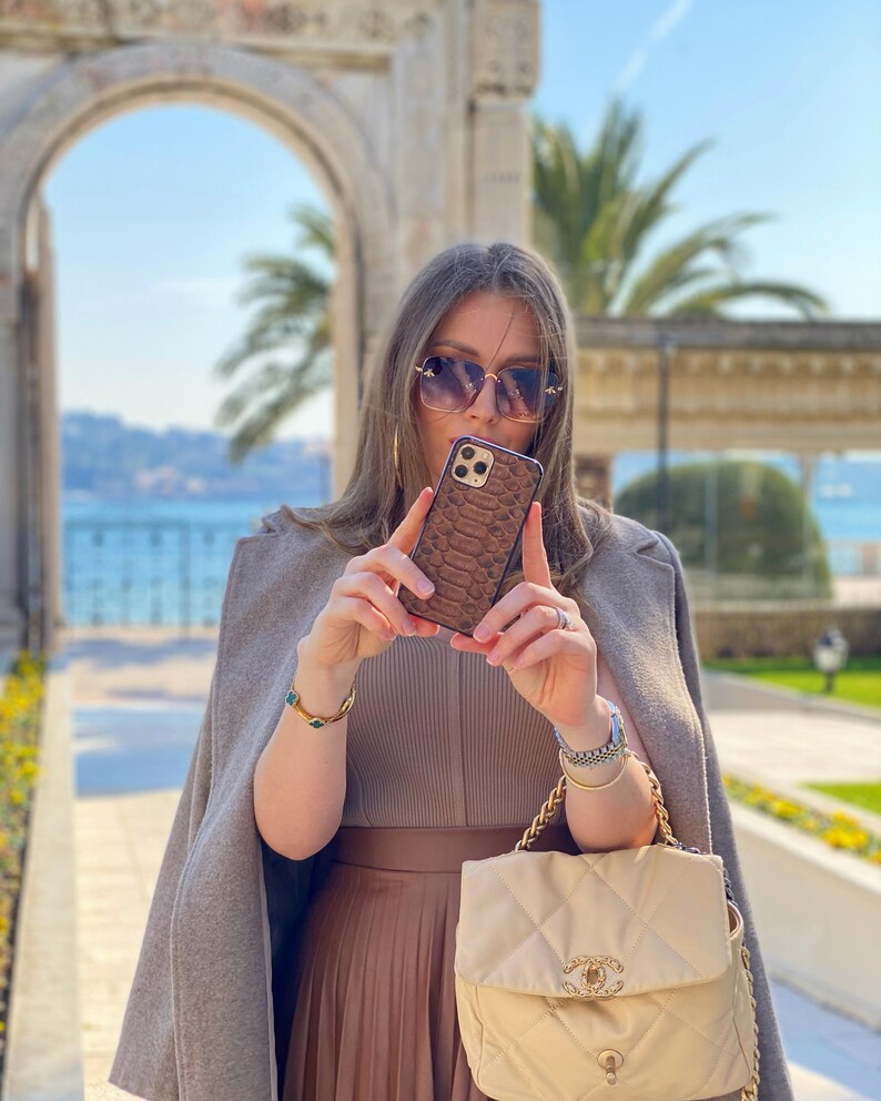 May include: A brown snakeskin-effect phone case is held up. The person is wearing sunglasses, a tan outfit, and carrying a cream quilted handbag with a gold chain strap. The background features a stone archway and palm trees, with a view of the sea.