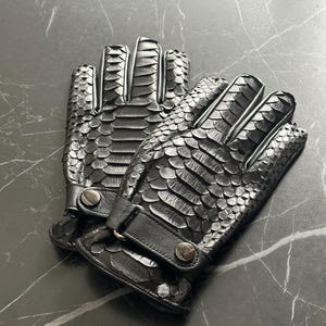 Könnte beinhalten: Ein Paar schwarze Lederhandschuhe mit Schlangenhautmuster. Die Handschuhe haben einen Handgelenksriemen und einen Knopfverschluss.