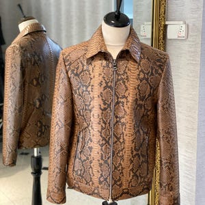 Puede incluir: Una chaqueta de cuero de serpiente marrón con cierre de cremallera. La chaqueta tiene cuello y dos bolsillos.