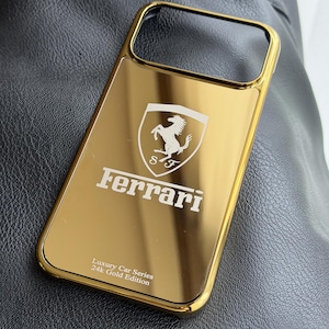 May include: A gold-coloured phone case featuring the Ferrari logo and text. The case has a reflective surface and the text "Luxury Car Series 24K Gold Edition". The Ferrari logo features a black prancing horse.