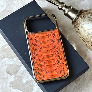 以下が含まれることがあります： オレンジ色のヘビ革模様のゴールドフレームの携帯電話ケース。ケースは濃い青色の箱の上に置かれ、背景には金の装飾品があります。ヘビ革模様には黒と茶色のアクセントがあります。