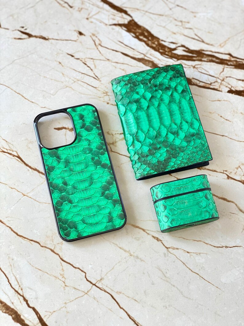 May include: A set of green snake-skin textured accessories, including a phone case, a wallet, and a small rectangular case, possibly for earbuds. The items are arranged on a marble surface.