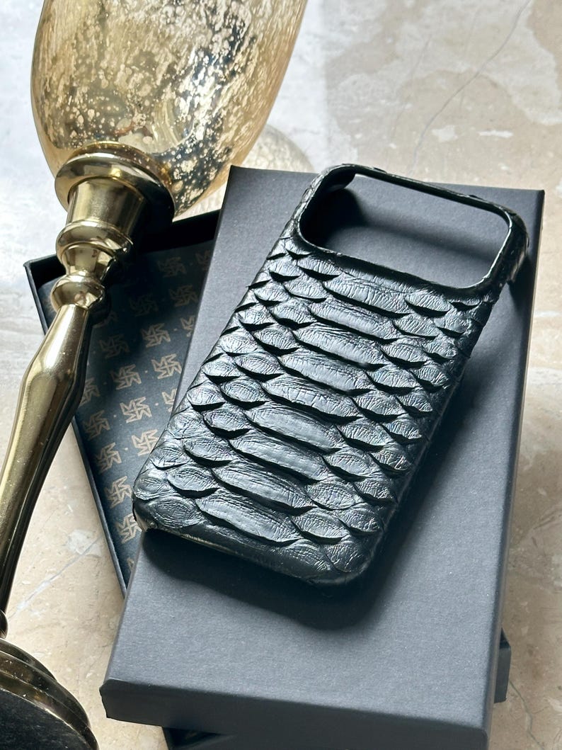 May include: A black phone case with a textured, scale-like pattern. The case is resting on a black box, with a gold-coloured decorative object in the background. The case appears to be made of leather.
