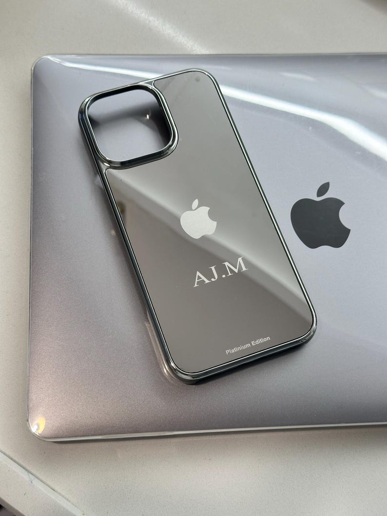 Puede incluir: Una funda transparente para tel&eacute;fono con marco plateado, apoyada sobre un port&aacute;til plateado. La funda tiene el logotipo de Apple y las iniciales "A.J.M" grabadas. Tambi&eacute;n es visible el texto "Platinum Edition".