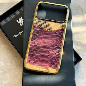 May include: A rectangular phone case featuring a polished gold-tone metal frame and a textured deep magenta snakeskin inlay. The top section has a gold-tone chevron pattern, and the bottom edge displays the text 24k Gold Python Edition.