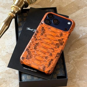 May include: An orange and brown snakeskin-effect phone case with a dark blue interior, presented in a black box with a thank you card. The website address www.mhiraje.com is visible on the card.