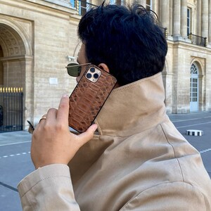 May include: A brown leather phone case with a reptile skin pattern. The case is held up to the ear. The background features a beige coat and a building with columns and windows.