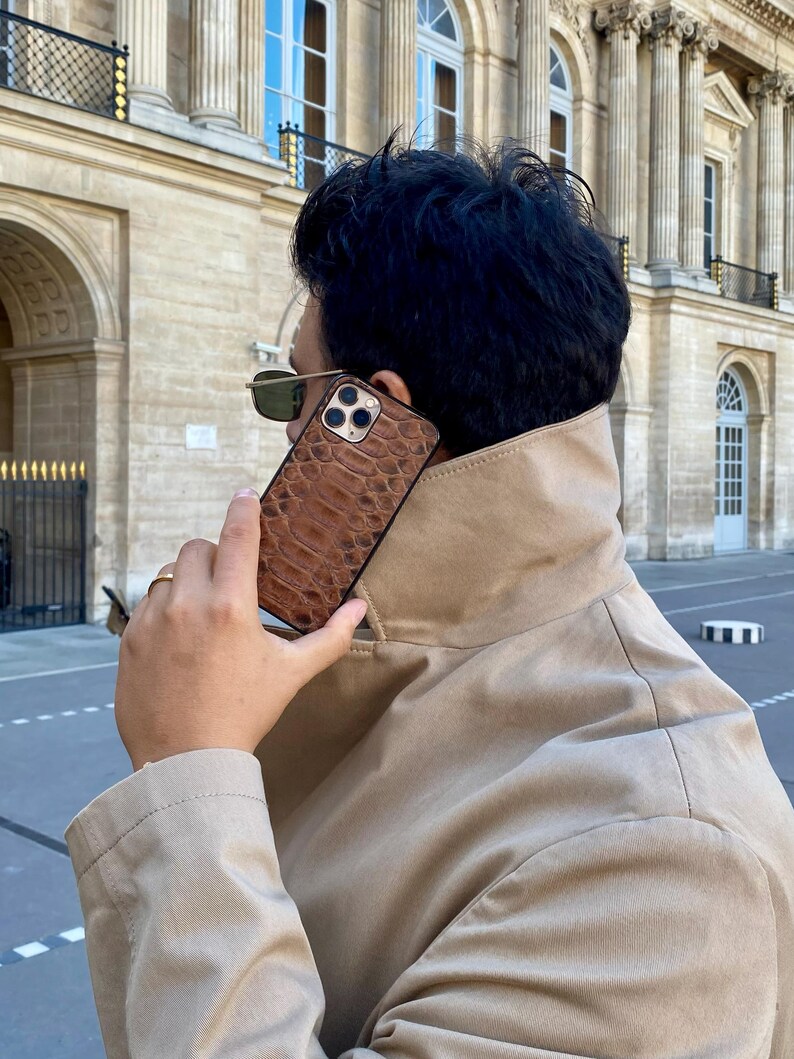 May include: A brown leather phone case with a reptile skin pattern. The case is held up to the ear. The background features a beige coat and a building with columns and windows.