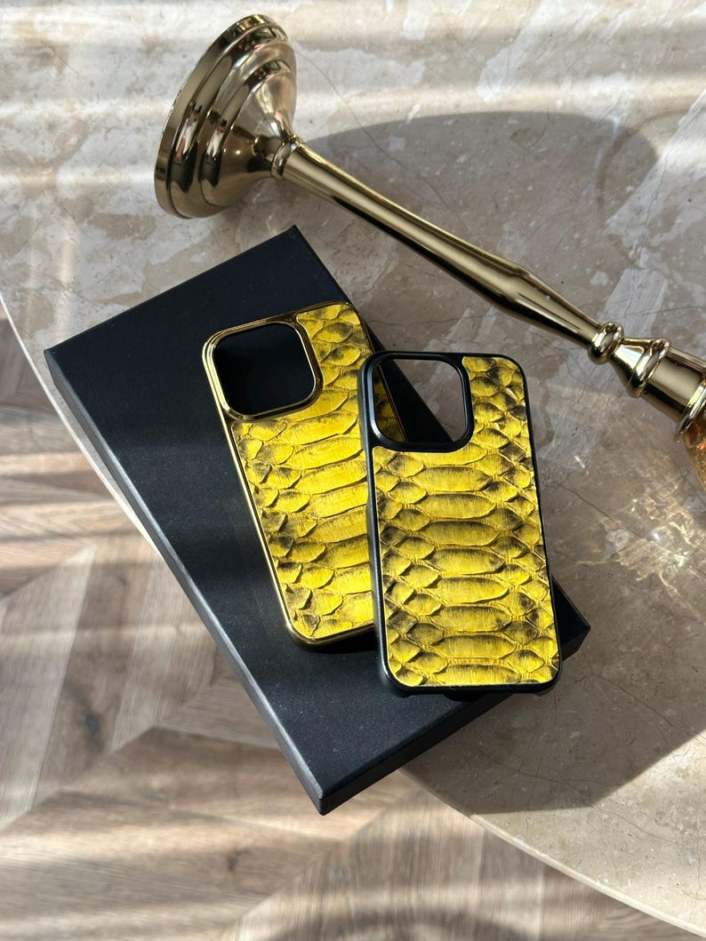 Pu&ograve; includere: Due custodie per telefono gialle e nere con un motivo a pelle di serpente sono esposte su una scatola nera. Un candelabro dorato &egrave; sullo sfondo. Le custodie per telefono hanno una cornice dorata.