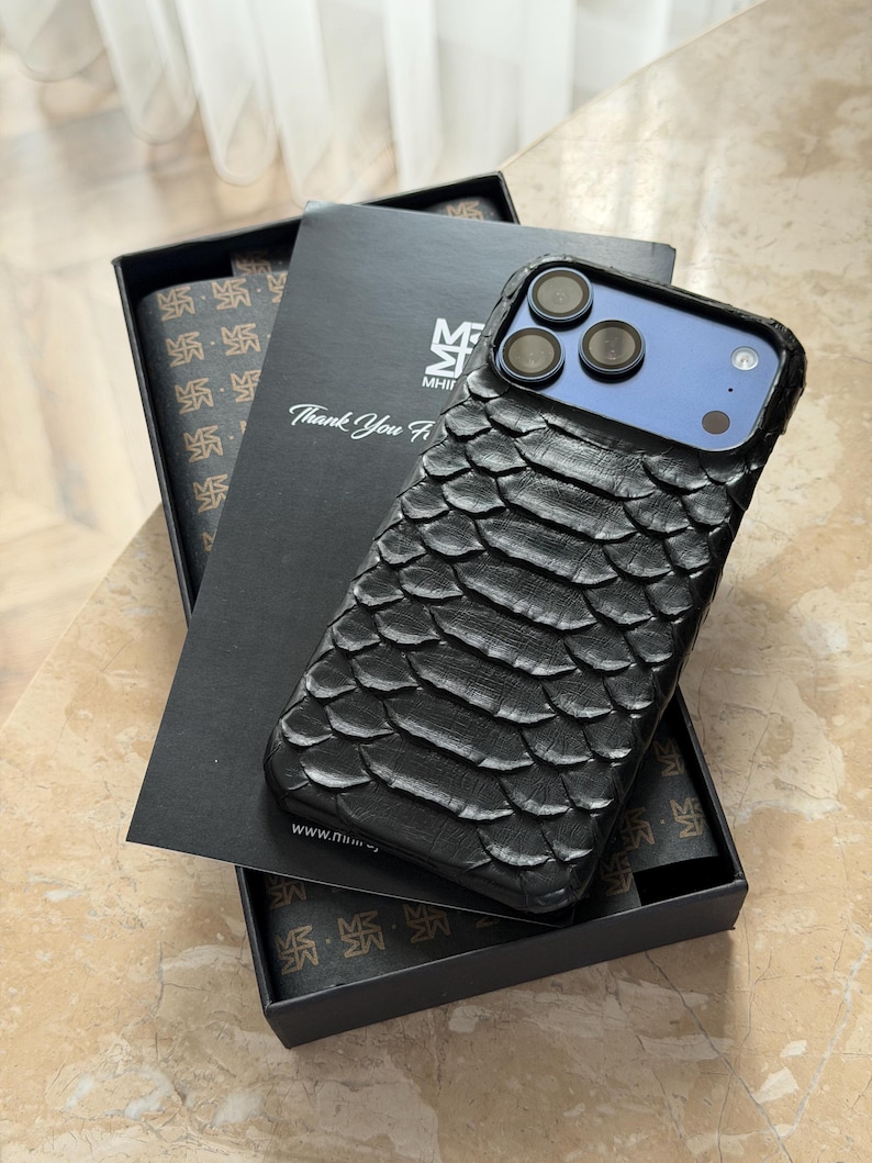 May include: Black textured phone case with a scale-like pattern, resting on a black box with the text "Thank You". The phone case has a blue camera module and is displayed with a matching patterned interior.