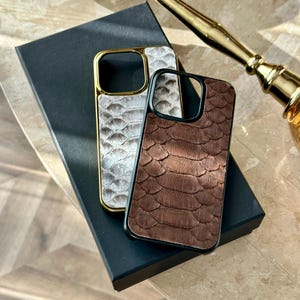 May include: Two phone cases with a reptile skin texture. One is white with a gold frame, the other is brown with a black frame. Both cases are on a black box, with a gold-coloured object in the background.