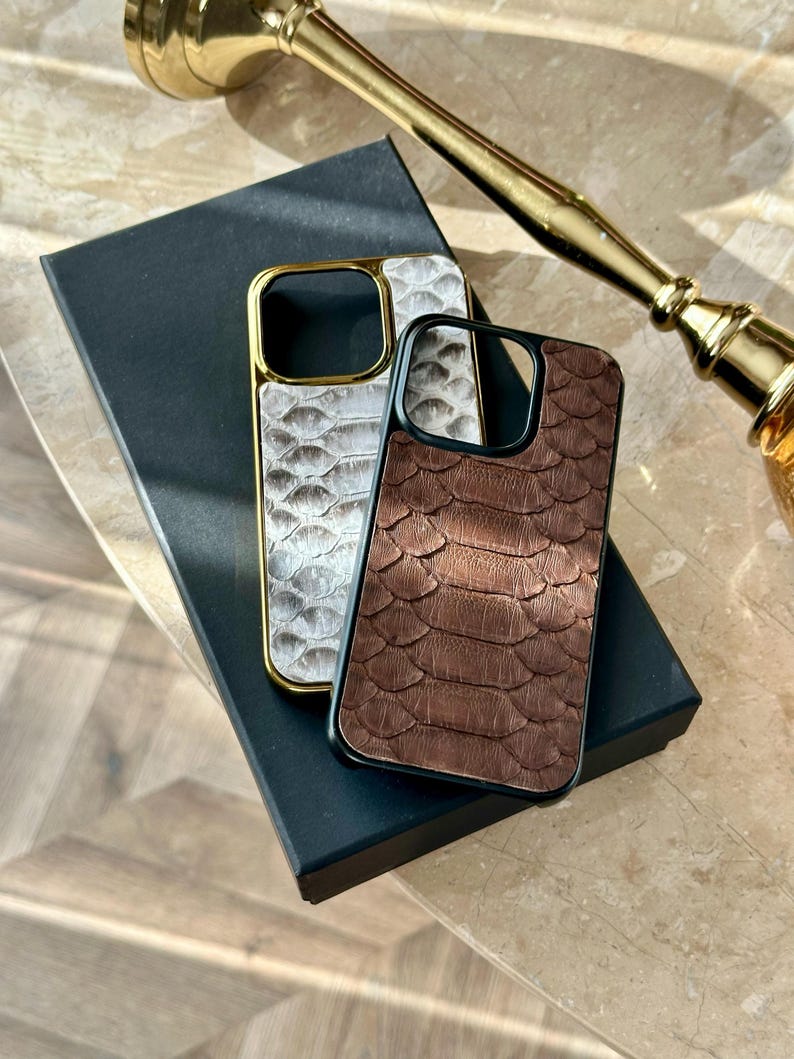 May include: Two phone cases with a reptile skin texture. One is white with a gold frame, the other is brown with a black frame. Both cases are on a black box, with a gold-coloured object in the background.
