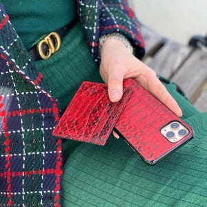 May include: Red textured leather phone case and matching wallet. The phone case is on a black frame. The wallet and phone case have a reptile skin pattern. The items are held in front of a green background.