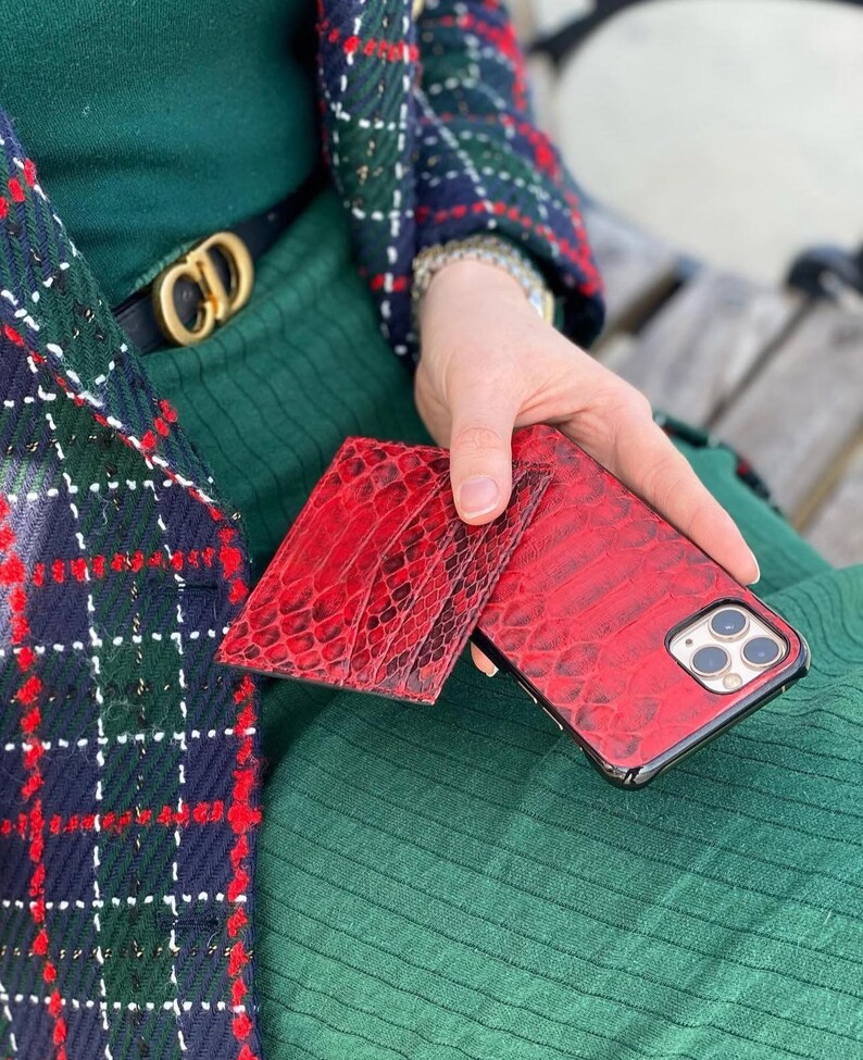 May include: Red textured leather phone case and matching wallet. The phone case is on a black frame. The wallet and phone case have a reptile skin pattern. The items are held in front of a green background.
