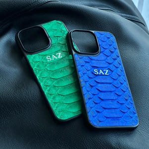 May include: Two phone cases with a textured snakeskin pattern. One is green, the other is blue. Both have a black border and the letters "SAZ" in the center. The cases are resting on a black leather surface.