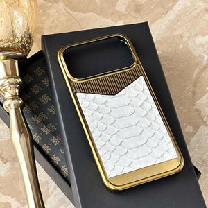May include: A gold-colored phone case with a white snakeskin-textured back and a gold frame. The case is resting on a black box. The words "24K Gold Plated Edition" are printed on the bottom of the case.