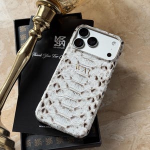May include: White and brown snakeskin-textured phone case with the initials "WAL" embossed on the back. The case is displayed in a black box with a gold-colored decorative object in the background. The case has a camera cutout.