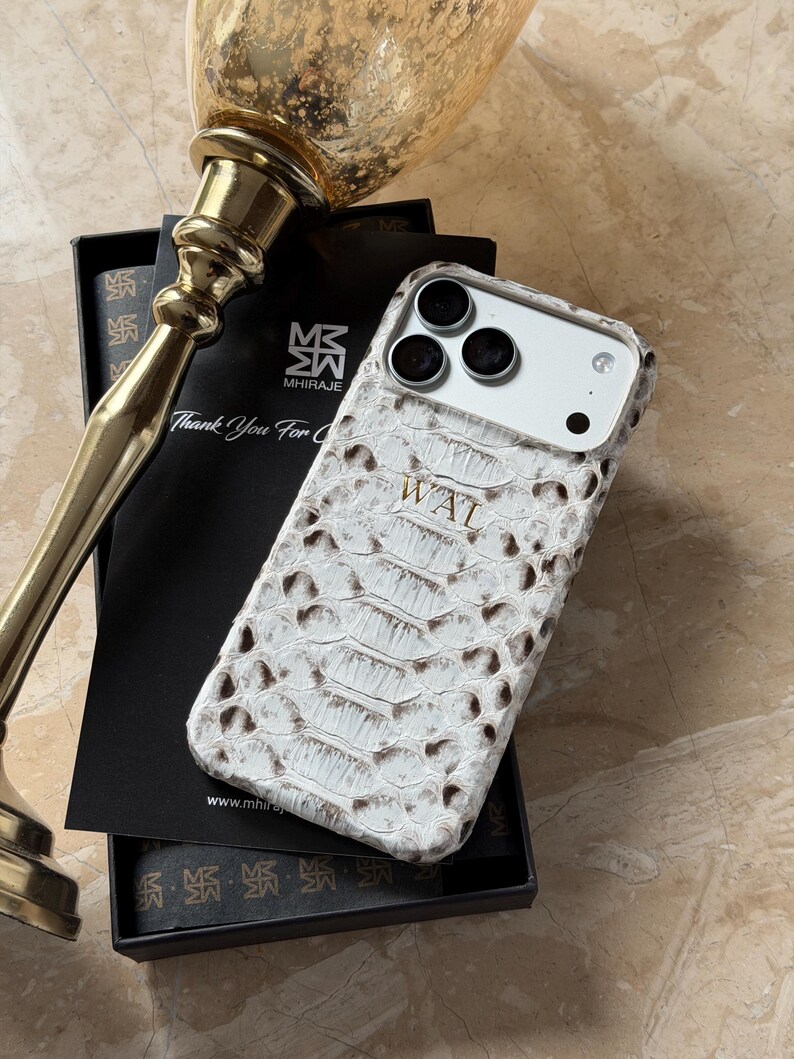 May include: White and brown snakeskin-textured phone case with the initials "WAL" embossed on the back. The case is displayed in a black box with a gold-colored decorative object in the background. The case has a camera cutout.