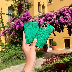 May include: A green snakeskin-textured phone case and matching wallet are held up against a backdrop of a yellow building and vibrant purple flowers. The phone case has a camera cutout. A gold and silver watch is visible on the wrist.