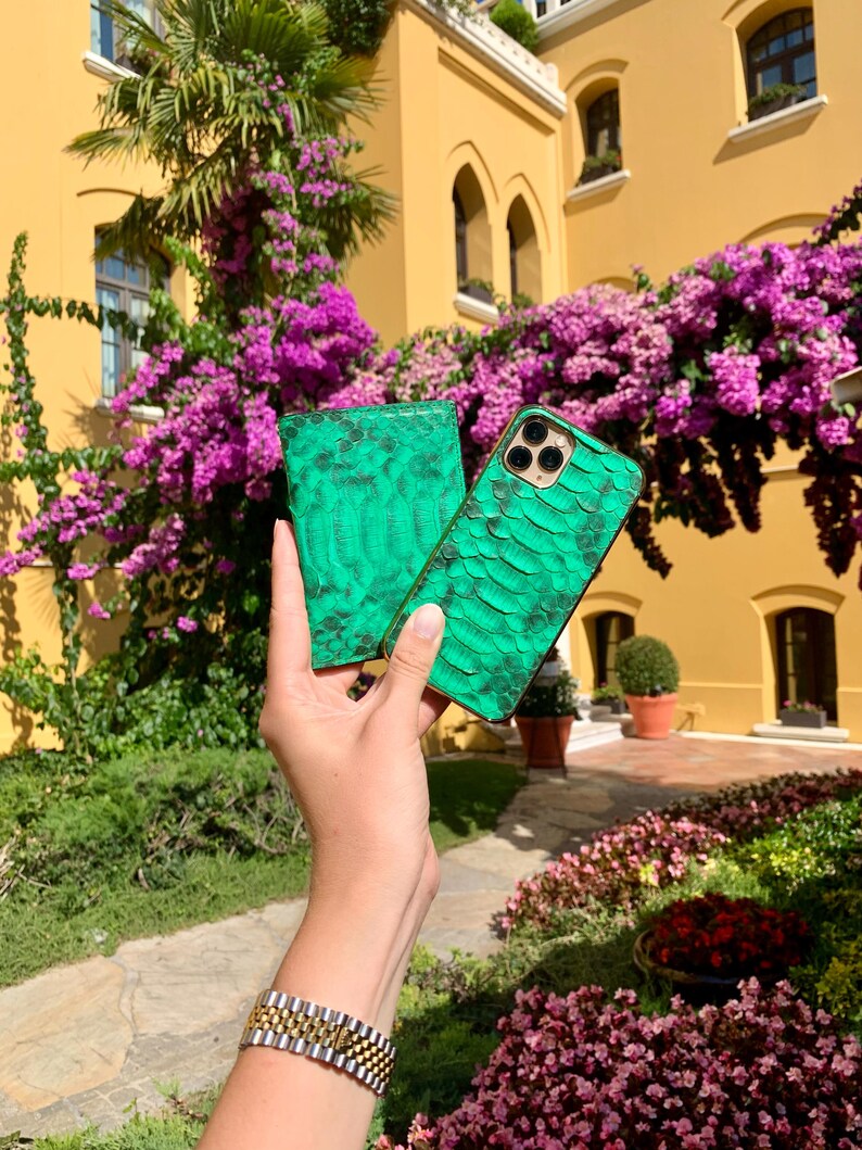 May include: A green snakeskin-textured phone case and matching wallet are held up against a backdrop of a yellow building and vibrant purple flowers. The phone case has a camera cutout. A gold and silver watch is visible on the wrist.