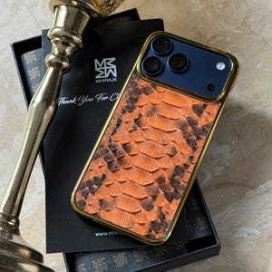 Peut inclure: Coque de smartphone avec un motif peau de serpent orange et un cadre doré. Le boîtier a une section supérieure bleu foncé avec trois objectifs d'appareil photo. Le boîtier est présenté sur une boîte noire avec le texte "Thank You For C" et la marque "MHIRAJE".