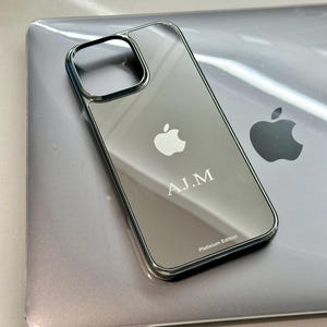 Puede incluir: Una funda plateada para iPhone con superficie reflectante, con el logotipo de Apple y las iniciales "A.J.M.". La funda tiene un borde met&aacute;lico y el texto "Platinum Edition". La funda est&aacute; sobre un port&aacute;til plateado.