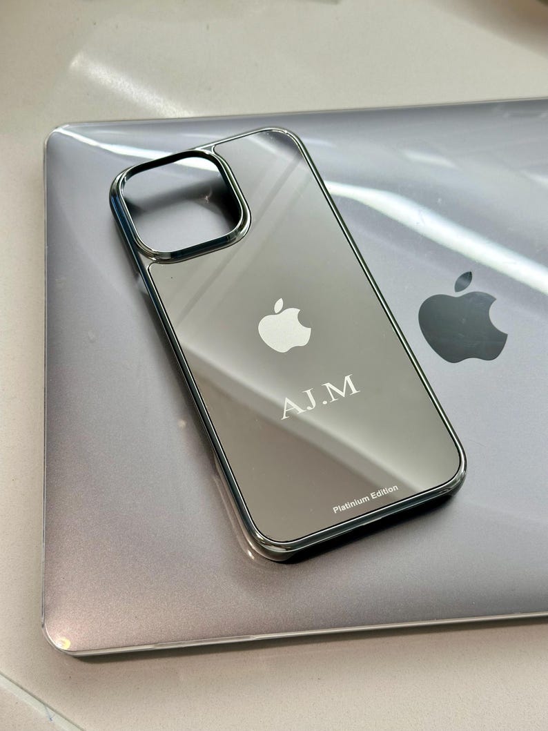 Puede incluir: Una funda plateada para iPhone con superficie reflectante, con el logotipo de Apple y las iniciales "A.J.M.". La funda tiene un borde met&aacute;lico y el texto "Platinum Edition". La funda est&aacute; sobre un port&aacute;til plateado.