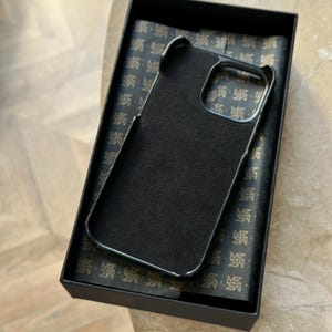 May include: A black phone case with a dark interior is presented in a black box. The case has a sleek, minimalist design with a smooth, matte finish. The box's interior features a patterned design. The case is designed to protect a phone.