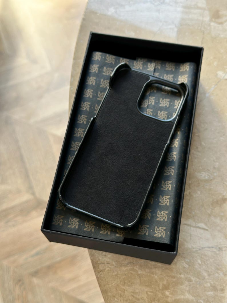 May include: A black phone case with a dark interior is presented in a black box. The case has a sleek, minimalist design with a smooth, matte finish. The box's interior features a patterned design. The case is designed to protect a phone.