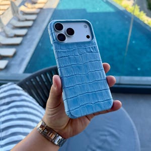 Peut inclure: Une coque de smartphone bleu clair avec un motif texturé en peau de crocodile. La coque présente une découpe rectangulaire pour le bloc photo, qui contient trois objectifs circulaires et un flash, révélant le dos blanc du téléphone.