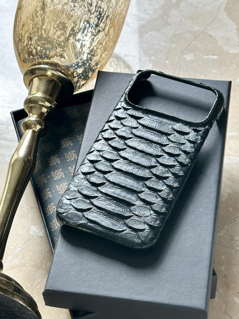 May include: Black phone case with a textured, scale-like pattern. The case is resting on a black box, with a gold-colored decorative object in the background. The case appears to be made of leather or a similar material.
