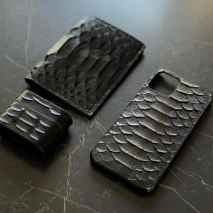 May include: A collection of black leather accessories: a wallet, phone case, and AirPod case. Each item features a textured, scale-like design. The accessories are presented on a dark, marble-effect surface.