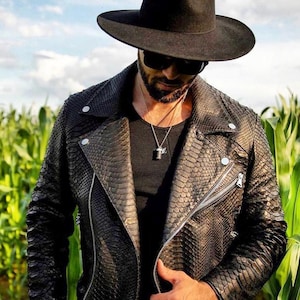May include: A man wearing a black leather jacket with a snakeskin pattern, a black fedora hat, and sunglasses. He is standing in a field of corn.