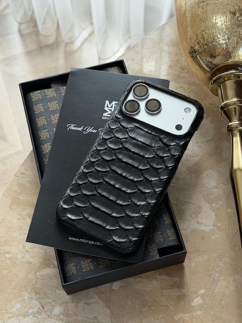 May include: Black textured phone case with a scale-like pattern, resting on a black box. The case has a camera cutout and is next to a box with the text "Thank You". The box has a repeating pattern and the website address www.mhiraje.com.