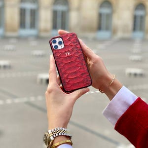 May include: Red textured phone case with the initials "TK" in white. The case has a snake-skin pattern and a black border. The phone case is held in front of a blurred background.